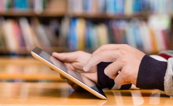 Tablet wird bedient, Hände im Close-Up, Bücherwand im Hintergrund unscharf