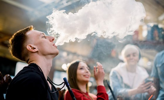 Gruppe junger Personen raucht Shisha. Mann im Vordergrund bläst Rauch aus.