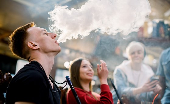Gruppe junger Personen raucht Shisha. Mann im Vordergrund bläst Rauch aus.