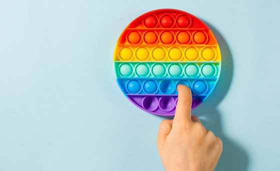 Rundes Fidget Toy in Regenbogenfarben liegt auf hellblauer Fläche. Ein Finger drückt Silikon-Bubble nach innen.