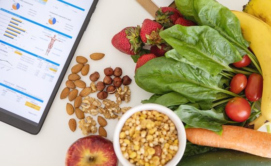 Von oben fotografiertes gesundes Essen, Nüsse, Bannanen, Erdbeeren, Salat, Tomaten liegen auf einer weiße, Untergrund, daneben ein Tablett auf dessen Display Diagramme, Zahlen abgebildet sind.