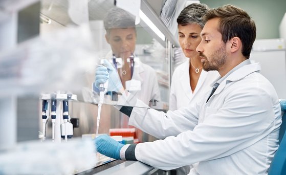 Zwei Wissenschaftler sind im Labor. Einer von ihnen pipettiert hinter einem Schutzglas. Wissenschaftlerin beobachtet ihn dabei.