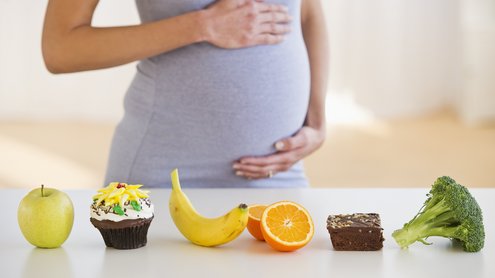Schwangere steht hinter einem Tisch und hält ihren Bauch. Auf dem Tisch steht ein Apfel, ein Mussif, eine Banane, eine Orange, ein Brownie und ein Brokkoli.