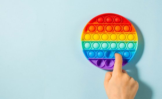 Rundes Fidget Toy in Regenbogenfarben liegt auf hellblauer Fläche. Ein Finger drückt Silikon-Bubble nach innen.