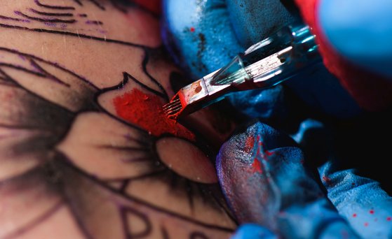 Tätowierfarbe Nahaufnahme von einer Hand, die einen blauen medizinischen Handschuh trägt und rote Farbe mit der Tätowiermaschine in Haut einarbeitet.